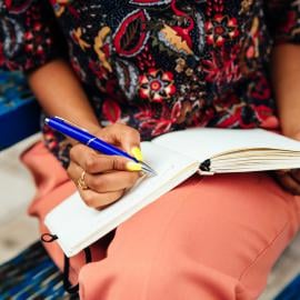 a girl sitting having a notepad on her lap and holding a pen 
