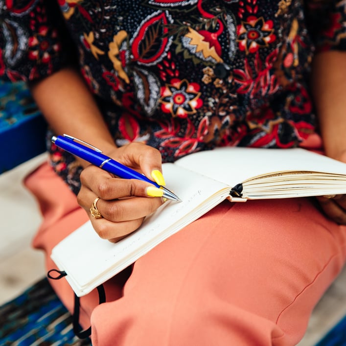 a girl sitting having a notepad on her lap and holding a pen 